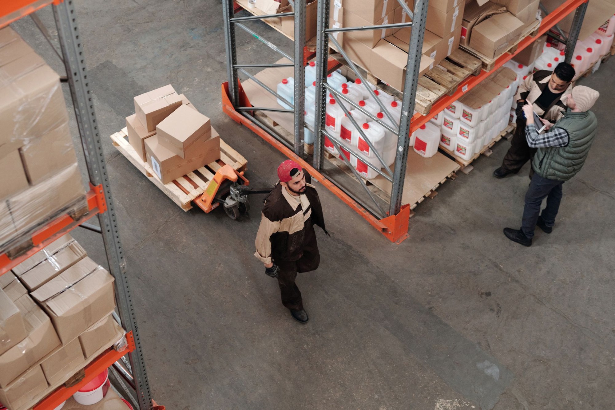 Three warehouse workers beside a pallet jack, cartons and stocked storage racks