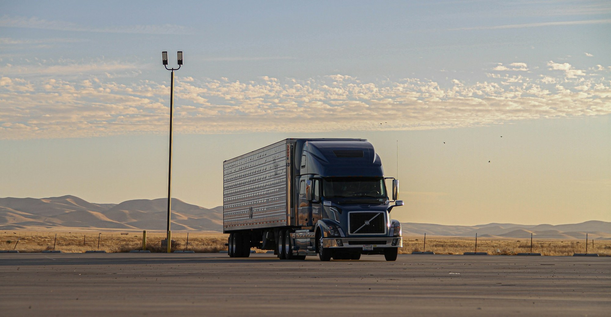 Dark blue semi truck and silver trailer on a paved lot beneath an evening sky